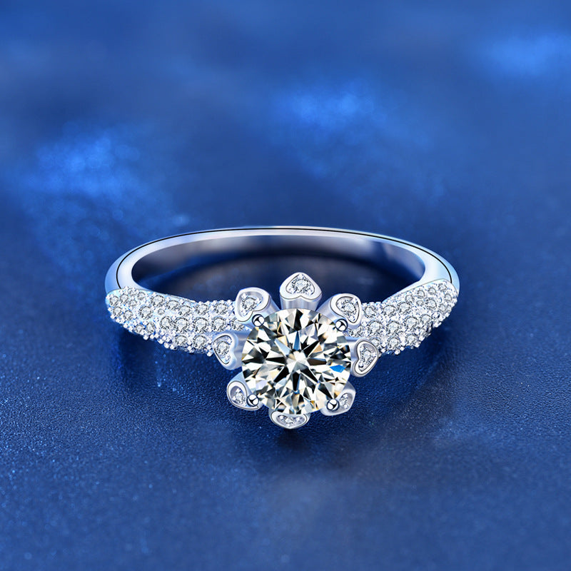 Top view of moissanite floral halo ring with heart accents and CZ pavé shoulders on blue background.