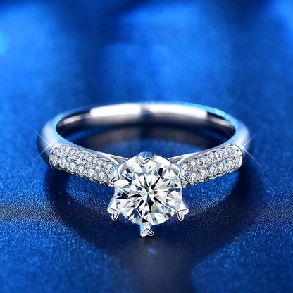 Six prong moissanite engagement ring with pavé CZ shoulders in sterling silver, front view on blue background.