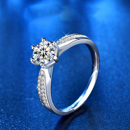 Side angle of 1 ct moissanite six-prong ring in sterling silver with pavé CZ shoulders on blue background