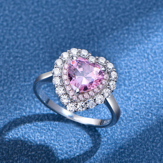 Pink heart-cut CZ double-halo ring in rhodium-plated sterling silver, angled view on blue background.